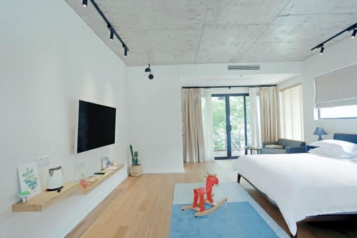 A modern hotel room with a large bed, a flat-screen TV, a wooden floor, a rocking horse, and a large window with curtains.
