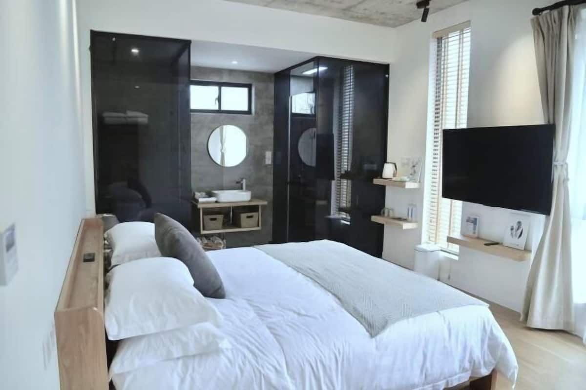 Un dormitorio con una cama, televisor, espejo y un baño visible a través de una puerta abierta.