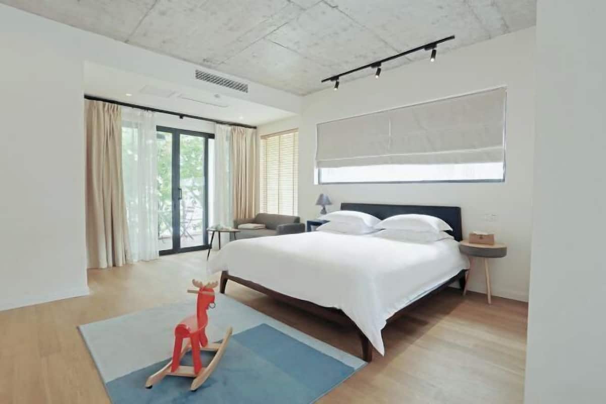 Una habitación de hotel moderna con una cama grande, una mesita auxiliar pequeña, una silla, una ventana con cortinas y un caballo balancín de madera.