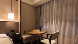 A hotel room with a bed, a desk, a chair, a small table with a lamp, and a view of the outside through curtains.