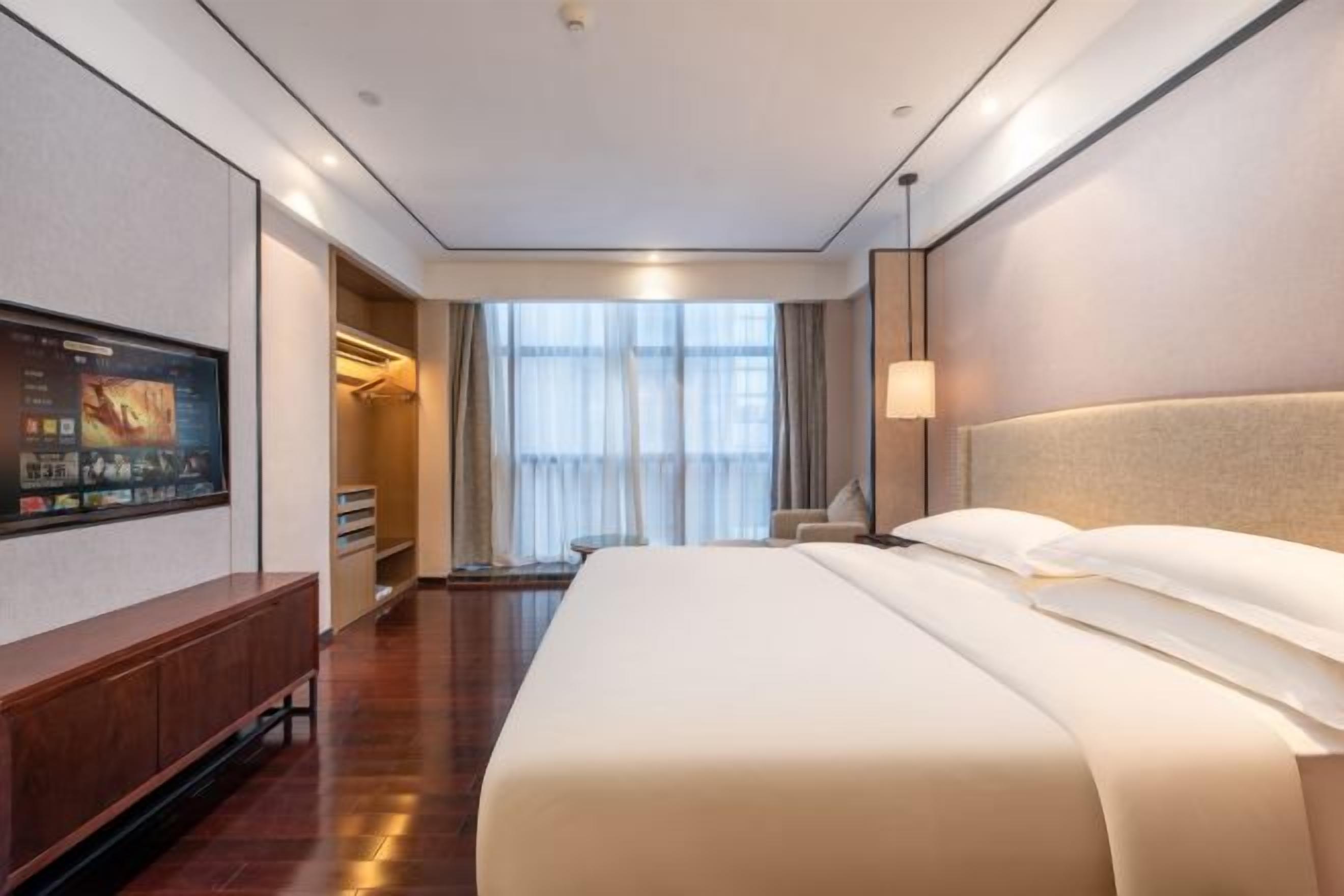 Une chambre d’hôtel dotée d’un grand lit, d’un téléviseur à écran plat fixé au mur, d’un coin salon avec un fauteuil et offrant une vue sur l’extérieur par la fenêtre.