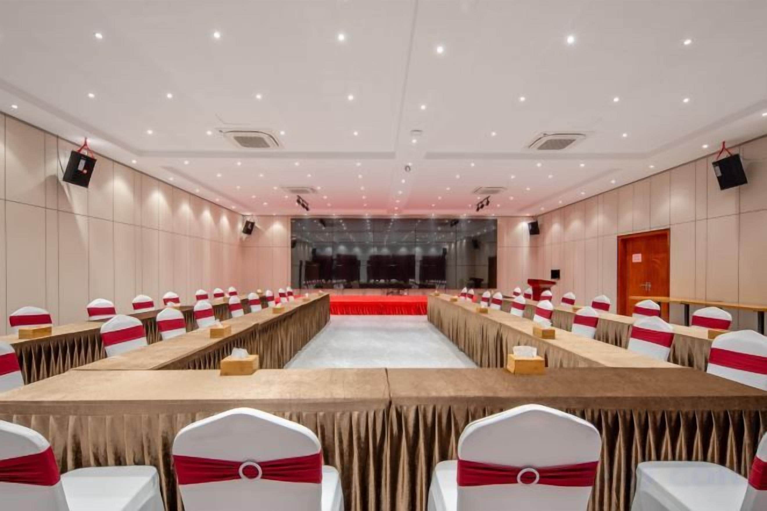 Una sala conferenze con lunghe tavolate, sedie, un palco e un grande schermo.
