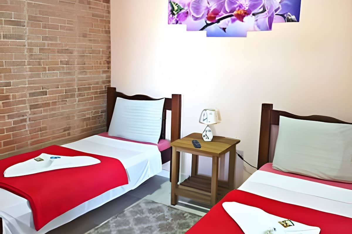 A hotel room with two single beds, each with a red and white bedspread, a small wooden nightstand with a lamp, and a brick wall.