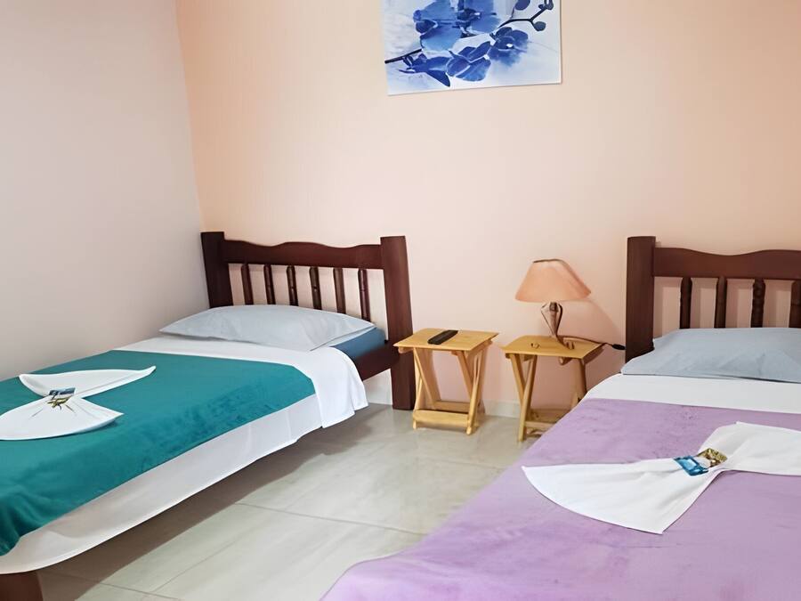 A hotel room with two single beds, each with a different colored bedspread and a towel with a logo. There are two bedside tables, one with a lamp and the other with a remote control.