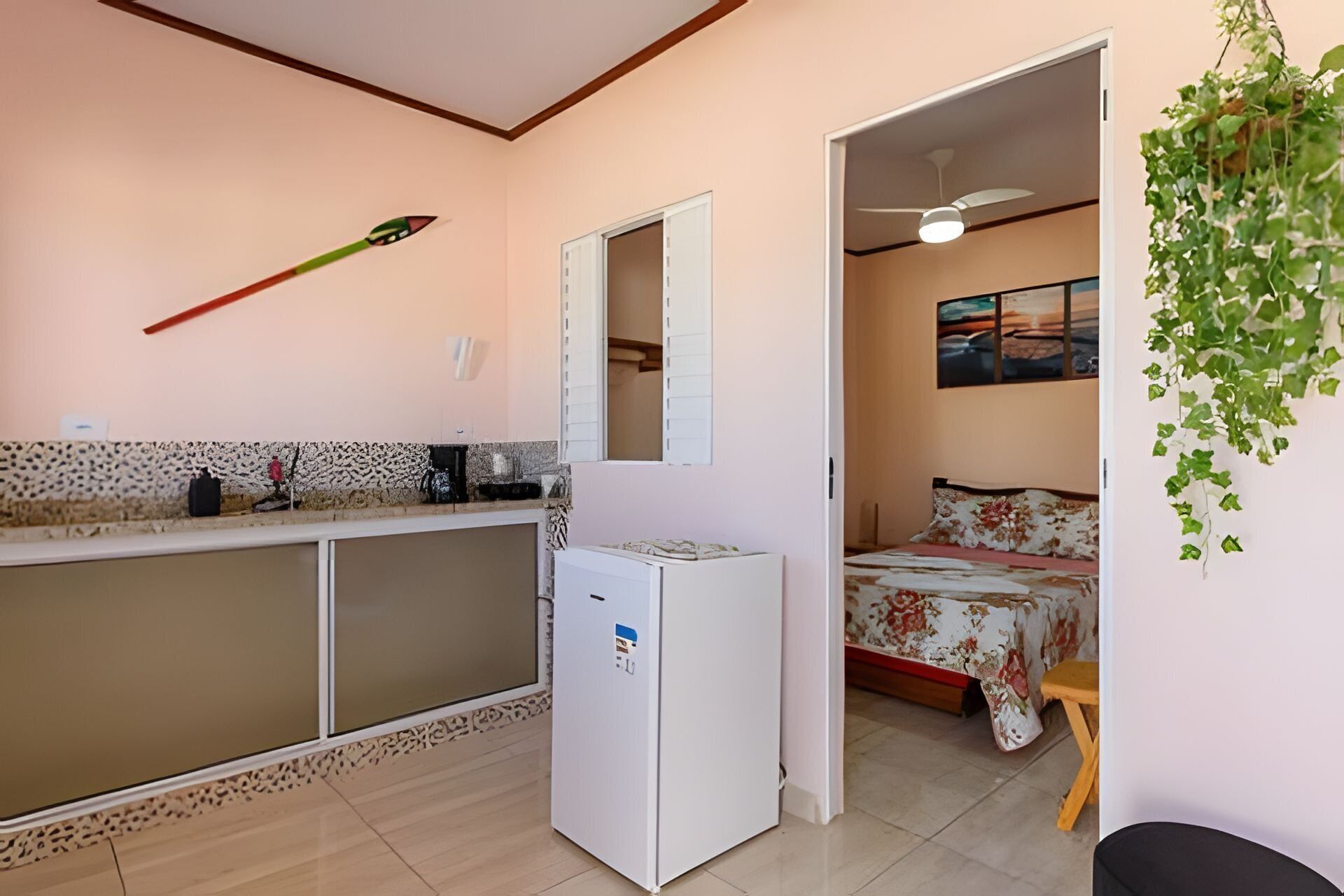 A compact kitchen with a refrigerator, a microwave, and a sink. There is a bed with a floral bedspread and a small wooden stool. A ceiling fan is visible, and a plant hangs from the ceiling.