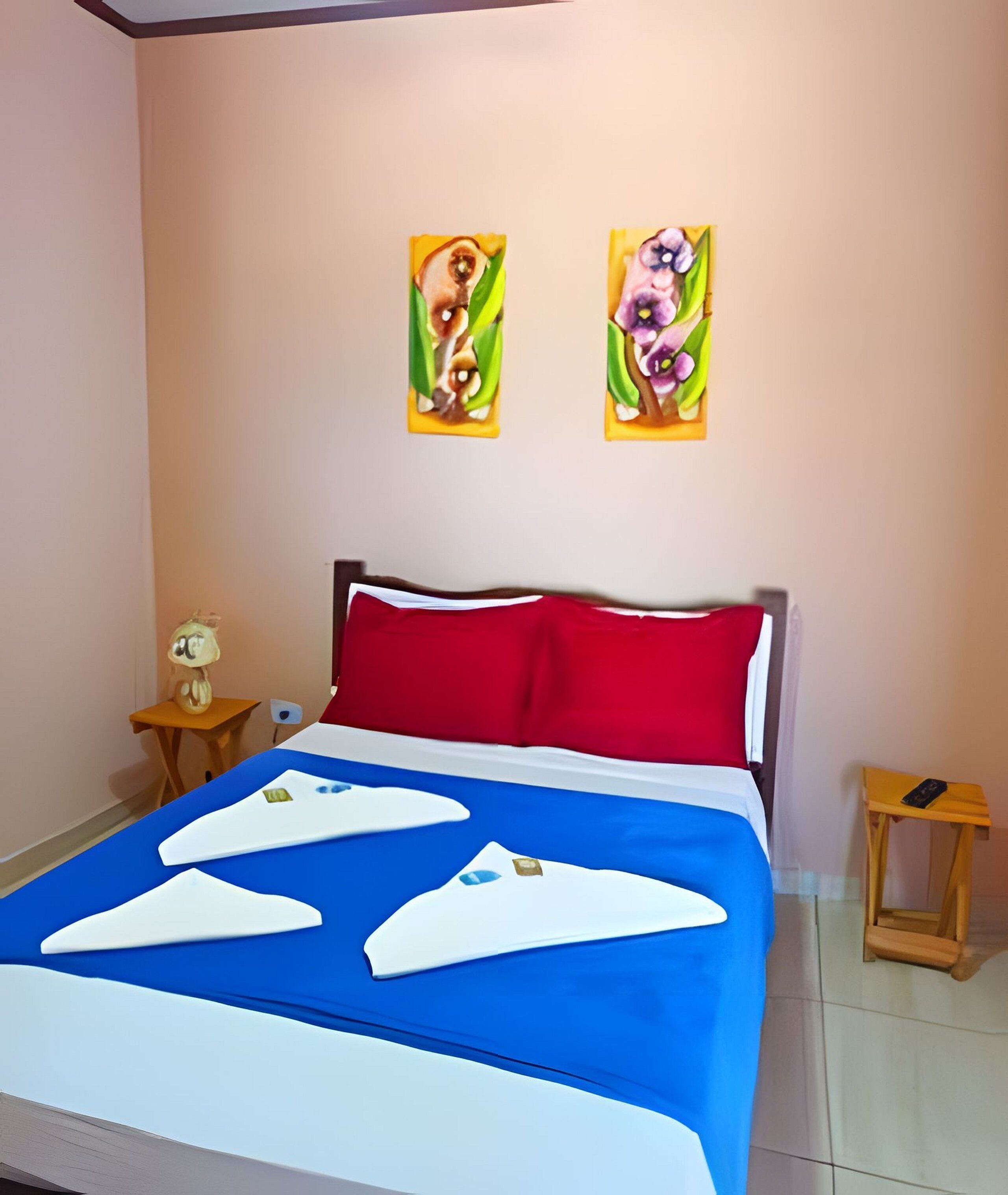 A hotel room with a bed, red and white pillows, a blue bedspread, two paintings on the wall, a small table with a lamp, and a wooden stool.