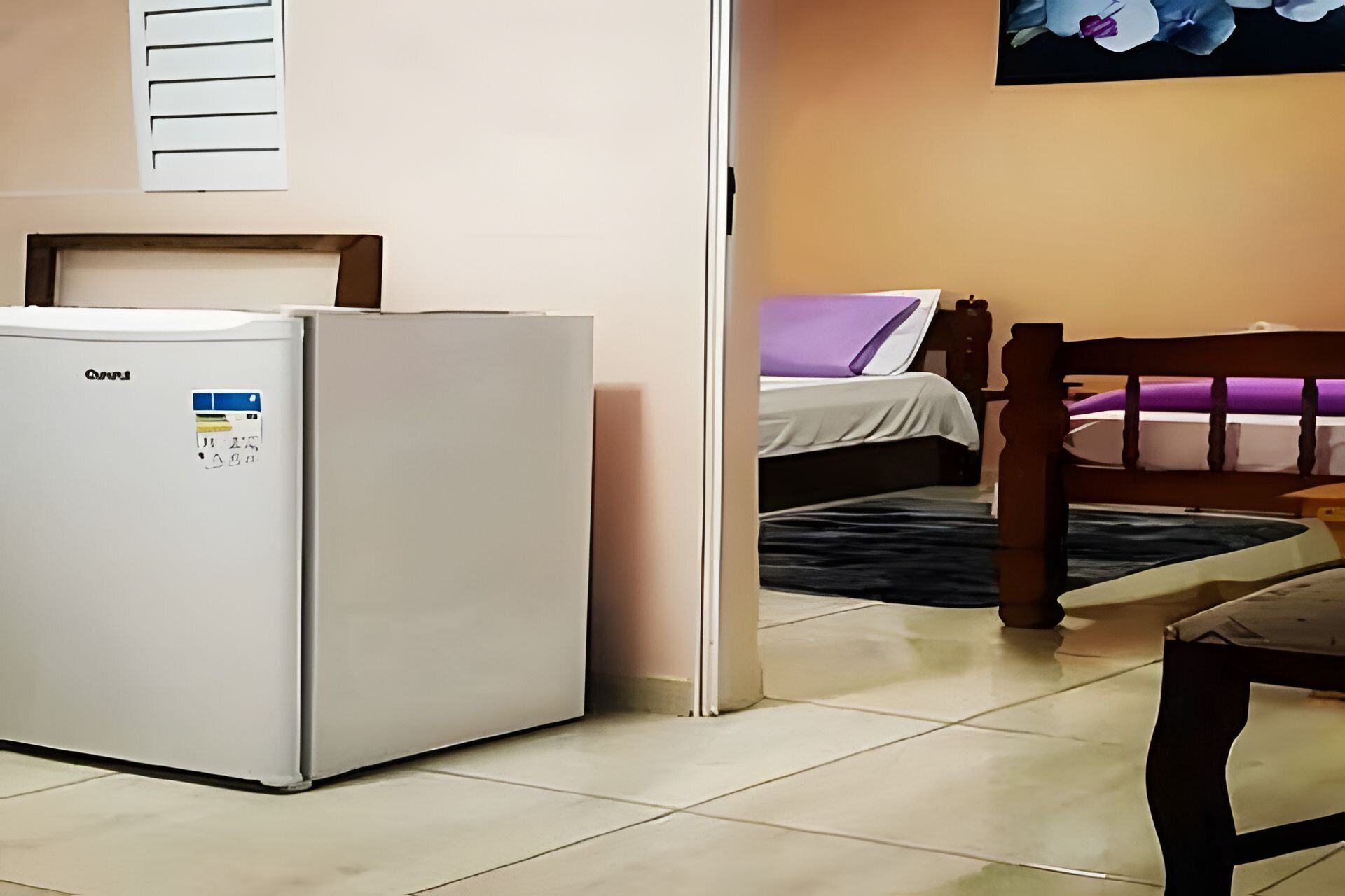 A compact room with a refrigerator, a bed with a purple pillow, and a wooden chair.