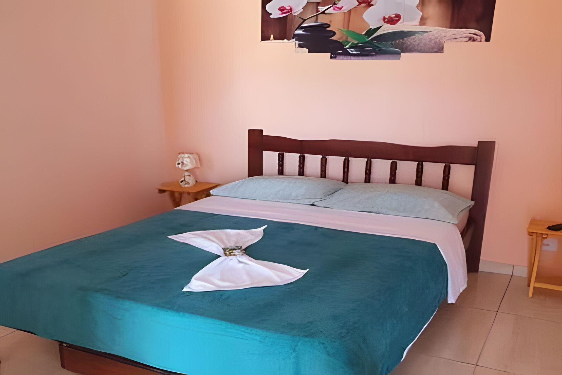 A neatly made bed with teal bedding and a white towel, a wooden headboard, a small bedside table with a lamp, and a wall-mounted picture of orchids.