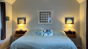 Une chambre à coucher comprenant un lit, deux tables de chevet, un banc, une œuvre d’art encadrée et une étagère fixée au mur.