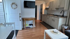 Une cuisine moderne avec des meubles en bois, un four encastré et un robinet noir. On y trouve une table à manger blanche avec un banc, une petite table d’appoint en bois et un canapé gris. Un téléviseur à écran plat est fixé au mur au-dessus de l’espace repas.