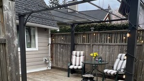 Une terrasse en bois avec une pergola, deux chaises à rayures, une petite table et un jardinier avec des fleurs.