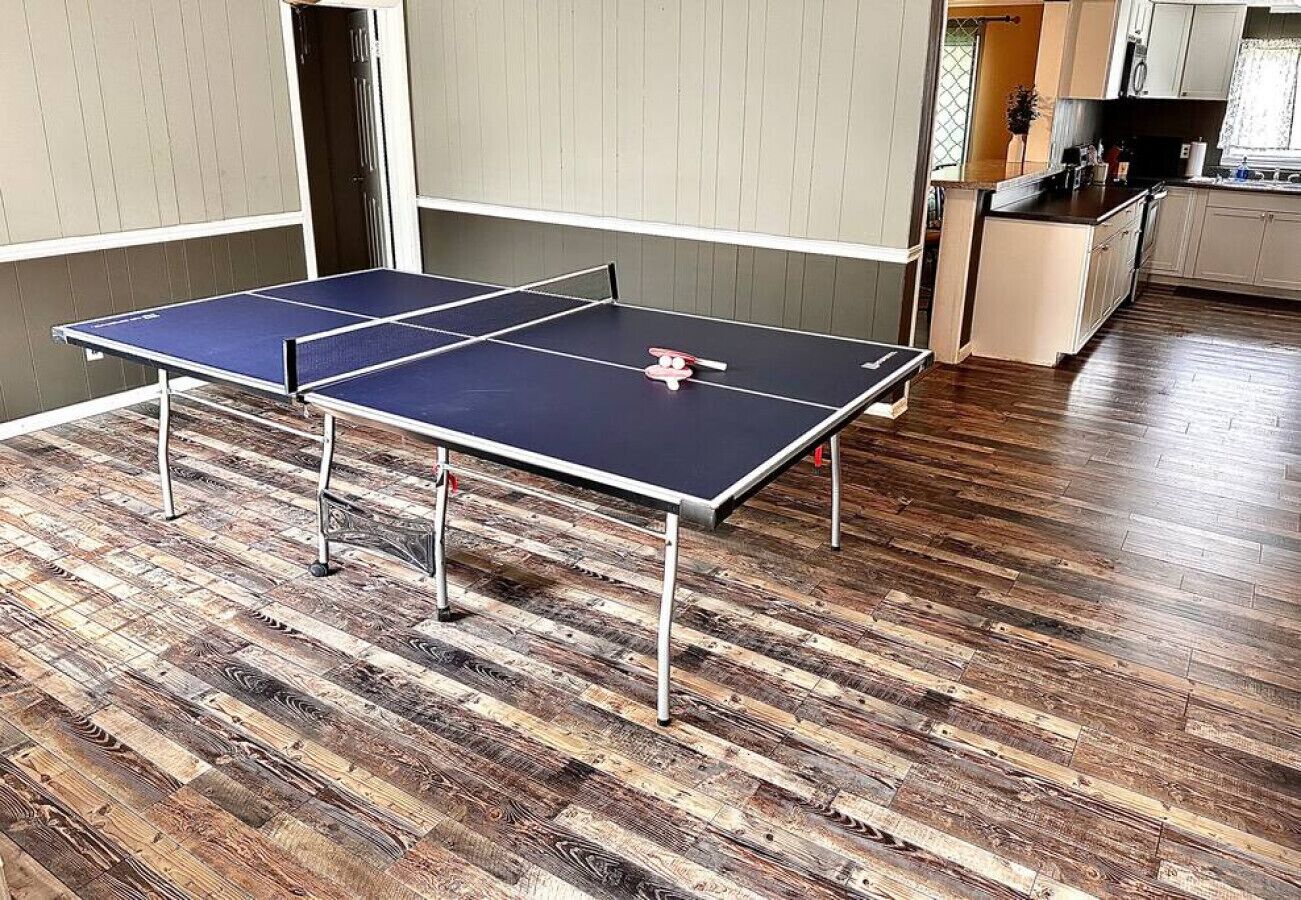 Una mesa de ping pong en una habitación con piso de madera y una cocina al fondo.
