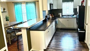Una cocina con piso de madera, gabinetes blancos, un refrigerador negro y una ventana con cortinas.