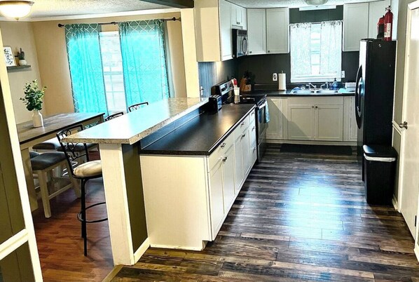 A kitchen with a wooden floor, white cabinets, a black refrigerator, and a window with curtains.