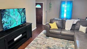 Una sala de estar con un televisor de pantalla plana sobre un soporte negro, un sofá de cuero marrón con cojines amarillos y blancos, y una alfombra estampada.