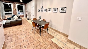 A modern living room with a sofa, dining table, and chairs. There are framed pictures on the wall and a window with curtains. - The Loft On Broad - Gadsden’s First Downtown Loft (Gadsden)