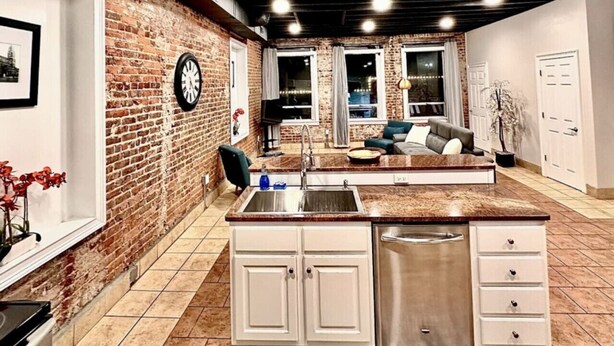 A kitchen with a brick wall, white cabinets, a stainless steel sink, and a dining area with a sofa. - The Loft On Broad - Gadsden’s First Downtown Loft (Gadsden)