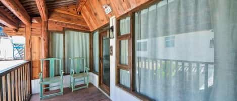 A wooden porch with a view of a building, two chairs, and a large window.