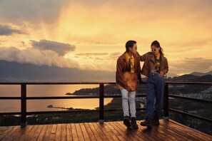A couple standing on a wooden deck overlooking a body of water with mountains in the background.