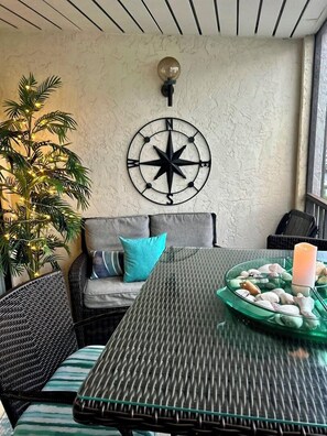 Un rincón al aire libre acogedor con una mesa redonda, sillas de mimbre, un sofá con cojines, una brújula decorativa en la pared y una planta en maceta.