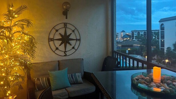 A balcony with a view of buildings, a compass wall decoration, a potted plant with lights, a sofa, and a glass table with candles. - Beautifully updated one bedroom one bath 4th-floor unit (Marco Island)