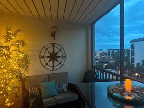 Ein Balkon mit Blick auf Gebäude, einer Kompass-Wanddekoration, einer Kübelpflanze mit Lichtern, einem Sofa und einem Glastisch mit Kerzen.