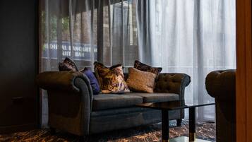 A dark-colored sofa with multiple cushions, a glass coffee table, and a view through sheer curtains.