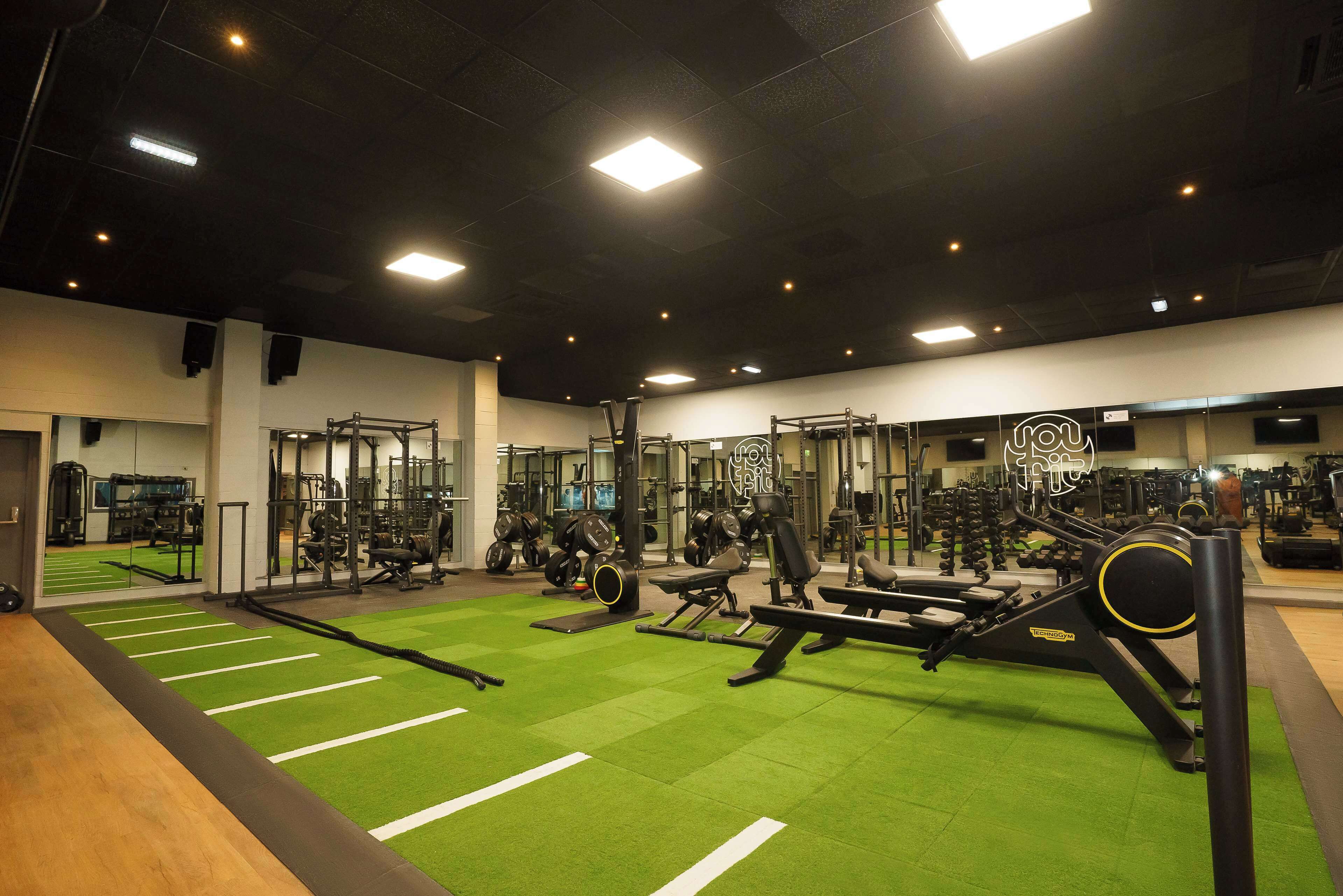 Un gimnasio con diferentes equipos de ejercicio, suelo verde y espejos en las paredes.