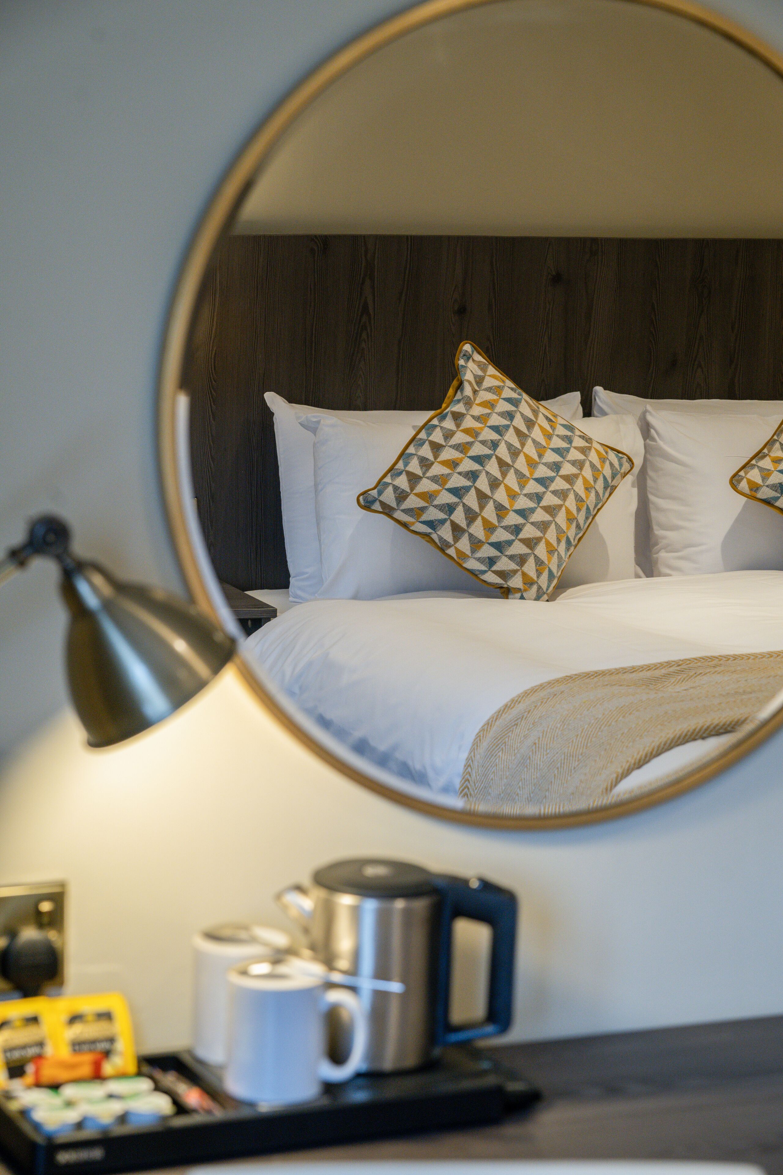 A hotel room with a bed, pillows, a bedside table with a lamp, a coffee maker, and a tray with toiletries.