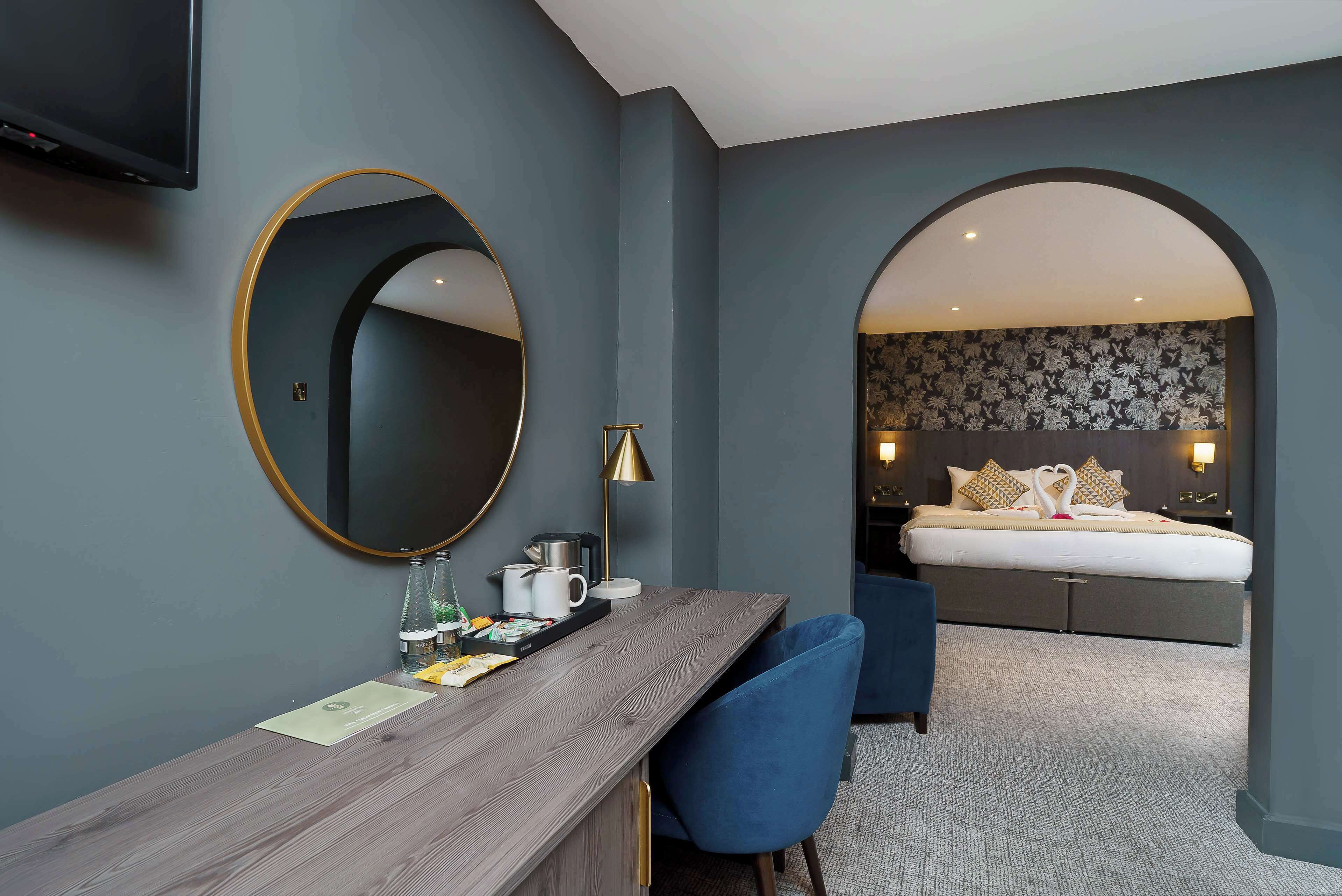 Una habitación de hotel con un escritorio de madera, un espejo redondo, una cama con cabecera floral y una silla azul.