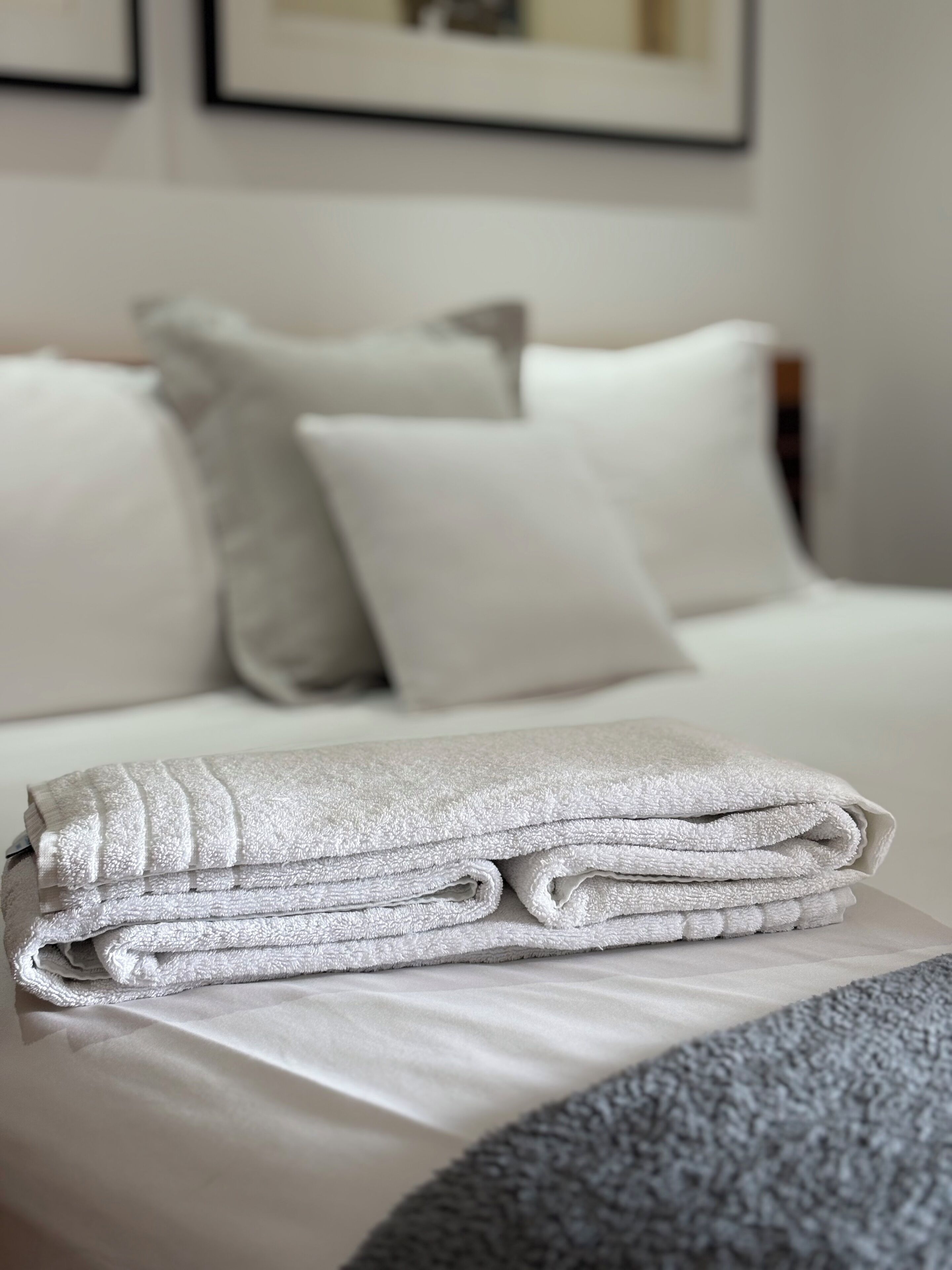 A neatly made bed with white linens and pillows, a folded towel on a bedside table, and a framed picture on the wall.