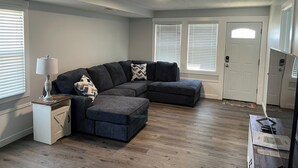 Una sala de estar moderna con un sofá modular, una mesita auxiliar y una lámpara. Hay dos ventanas con persianas, una puerta y una luz de techo.