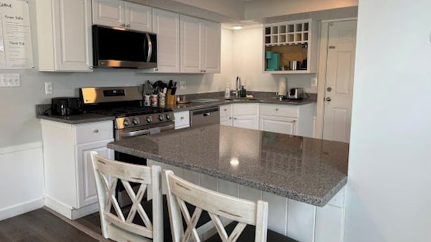 Una cocina con armarios blancos, encimera de granito, microondas y estufa. Hay dos sillas de madera con respaldo de listones cruzados.