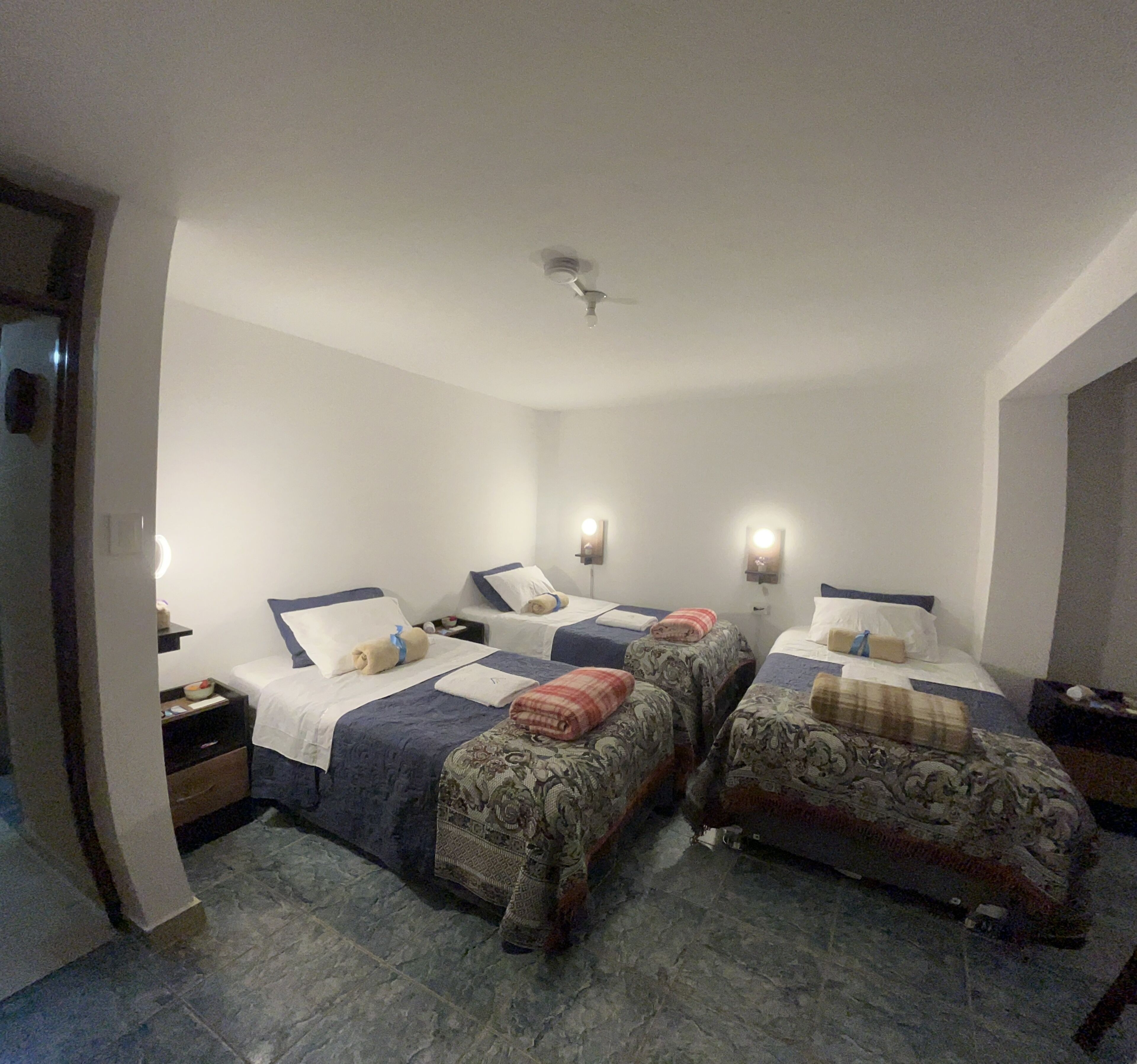 A hotel room with two beds, each with a pillow and a blanket. There are bedside tables with lamps and a ceiling fan.