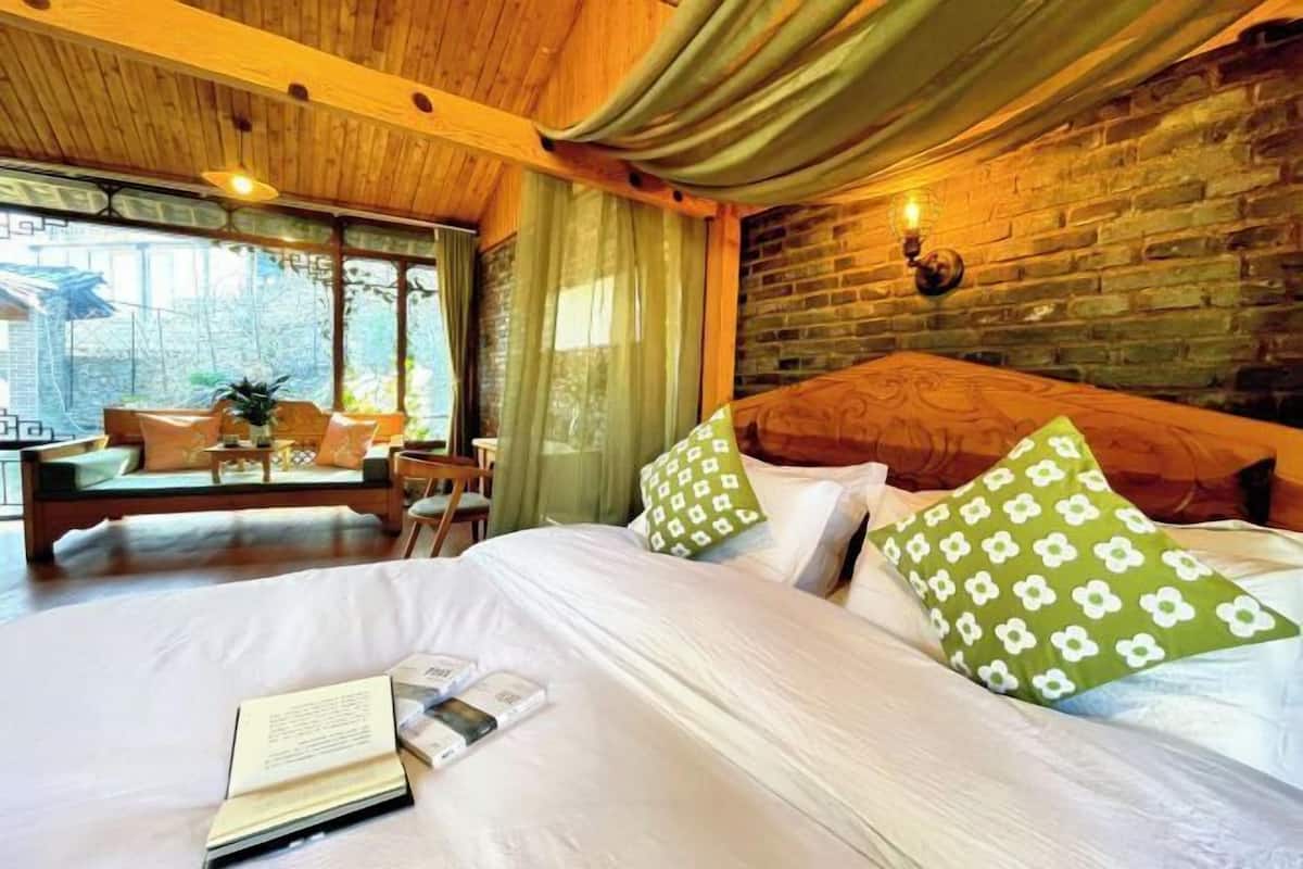 A hotel room with a wooden ceiling, a brick wall, a large bed with white bedding and green patterned pillows, a small table with a book, and a window with curtains.