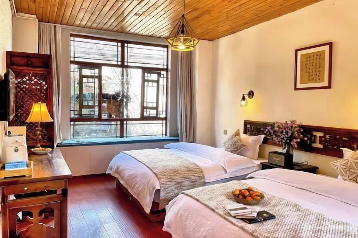 A hotel room with two beds, a wooden ceiling, a large window with curtains, a bedside table with a lamp, and a wall-mounted light fixture.