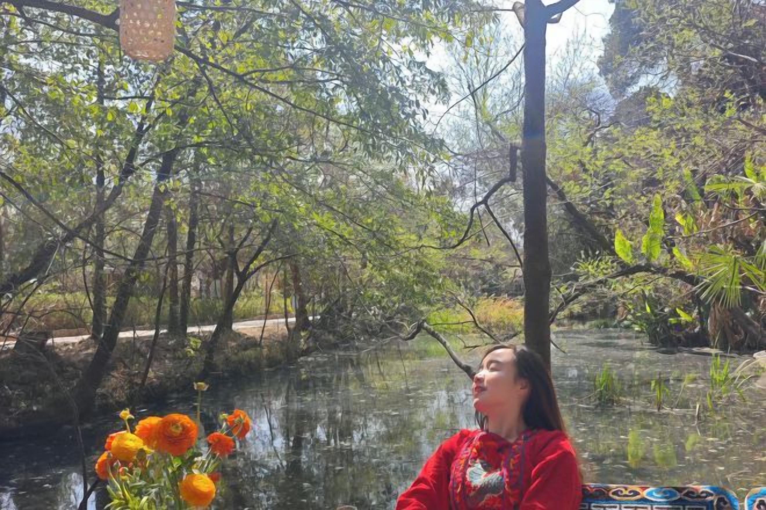A person sitting by a body of water with trees and a wooden structure in the background.