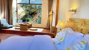 Une chambre d’hôtel avec un grand lit, un coin salon avec une table et des chaises, une vue sur la verdure par la fenêtre et une tête de lit en bois.