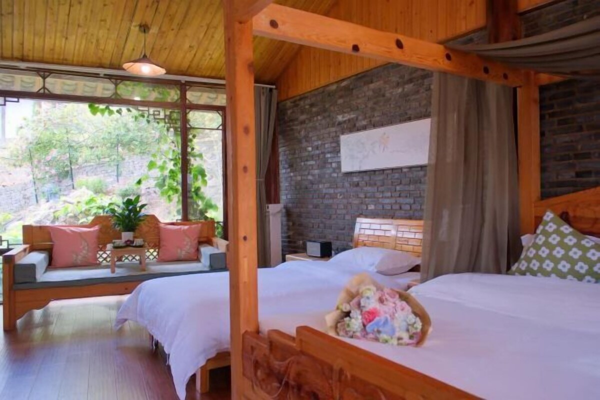 A wooden four-poster bed with a teddy bear, a sofa with cushions, and a view of greenery outside.