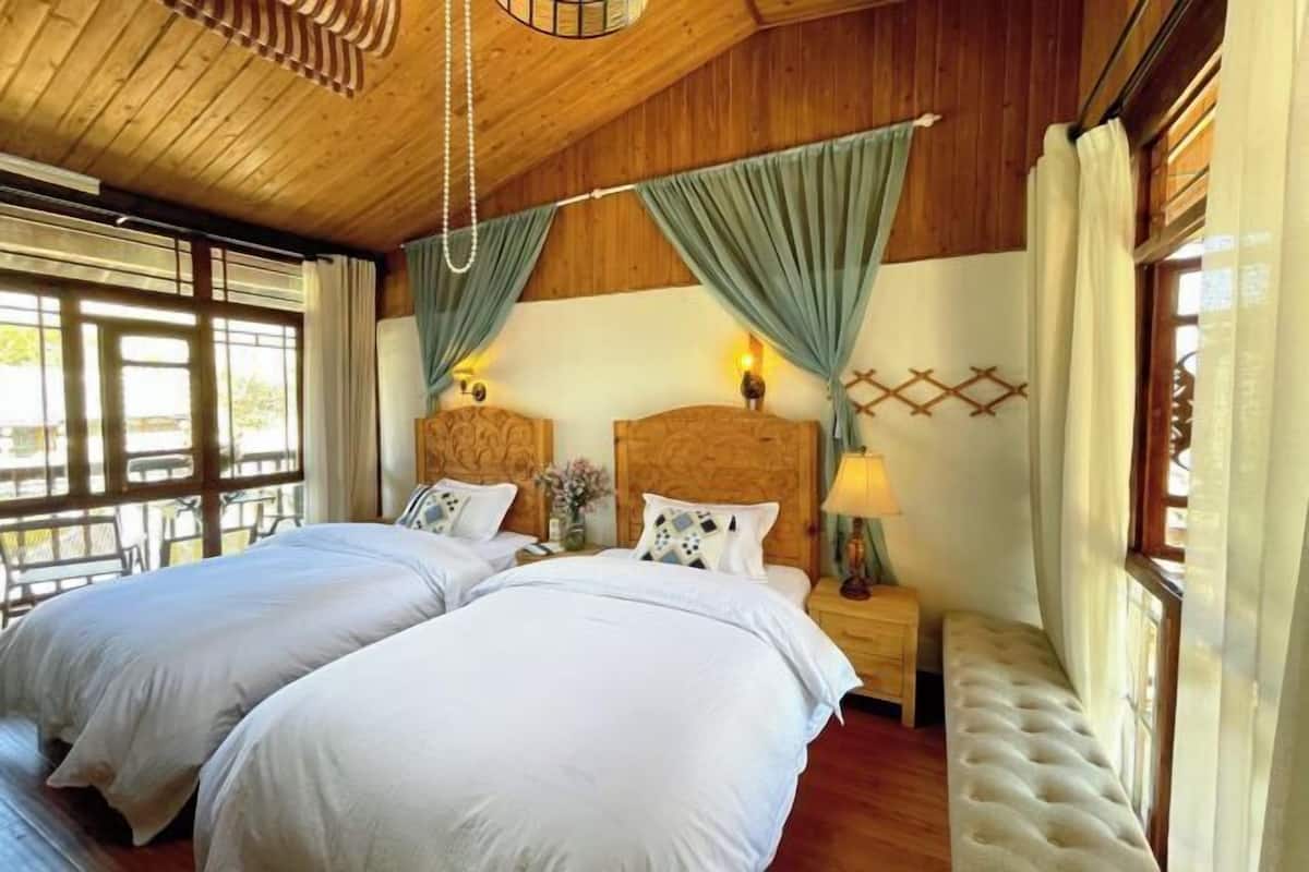 A room with two beds, wooden ceiling, curtains, bedside lamps, and a bench.
