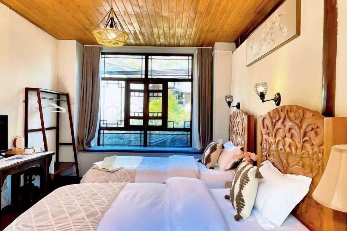 A hotel room with a wooden ceiling, a large window with curtains, a bed with patterned bedding, a bedside table with a lamp, and a wall-mounted TV.