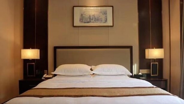 A hotel room with a large bed, two bedside lamps, a framed picture on the wall, and a wooden headboard. - Shizhushan dezheng Hotel (Yizheng)