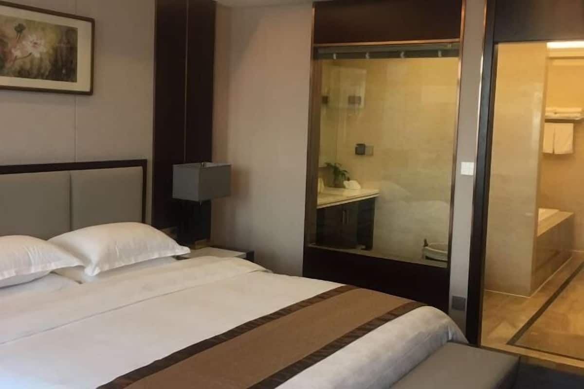 A hotel room with a large bed, a bedside table with a lamp, and a view of the bathroom through a glass door.