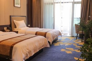 Habitación de hotel con dos camas, un ventanal con cortinas, una planta y una alfombra azul con estampado floral.