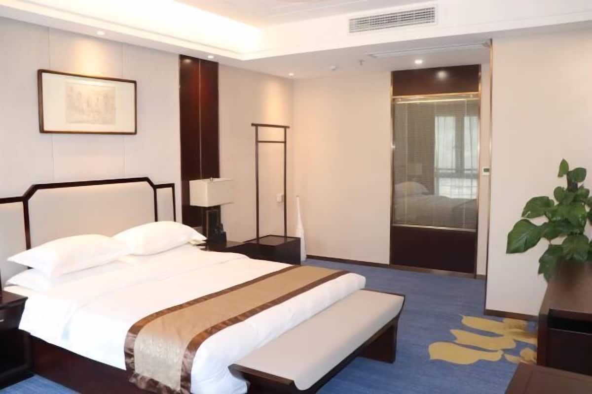 A hotel room with a large bed, bedside tables, a bench, a floor lamp, a potted plant, and a door leading to another room.