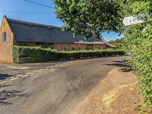 En landevej der fører til en murbygning med stråtak, omgivet af grøn vegetation og en klar himmel.