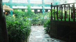 A courtyard with a tiled path, lush greenery, and a thatched-roof structure.