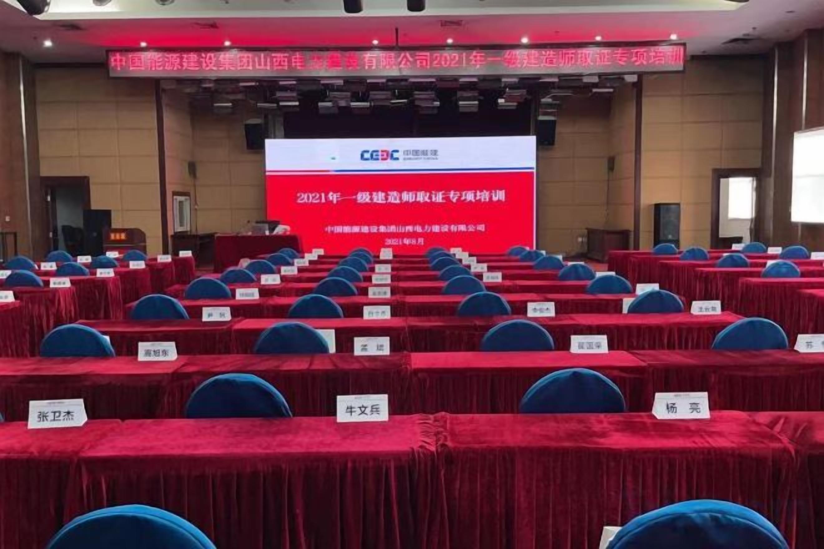 Un salón de conferencias con filas de mesas y sillas, una pantalla grande mostrando texto, y un escenario con un atril.
