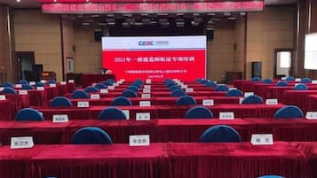 Un salón de conferencias con filas de mesas y sillas, una pantalla grande mostrando texto, y un escenario con un atril.