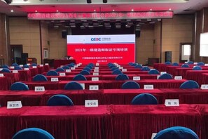 Un salón de conferencias con filas de mesas y sillas, una pantalla grande mostrando texto, y un escenario con un atril.